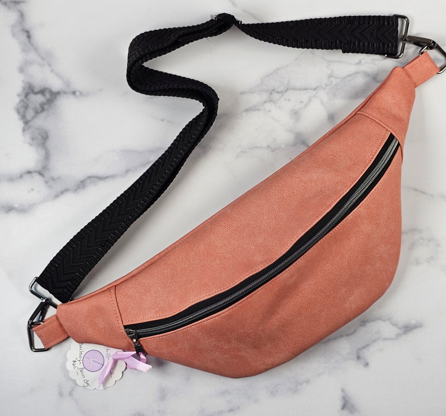 Pink faux leather sling bag with black accents displayed on a table, showing its sleek design and Rifle Paper Co. book-pattern interior.