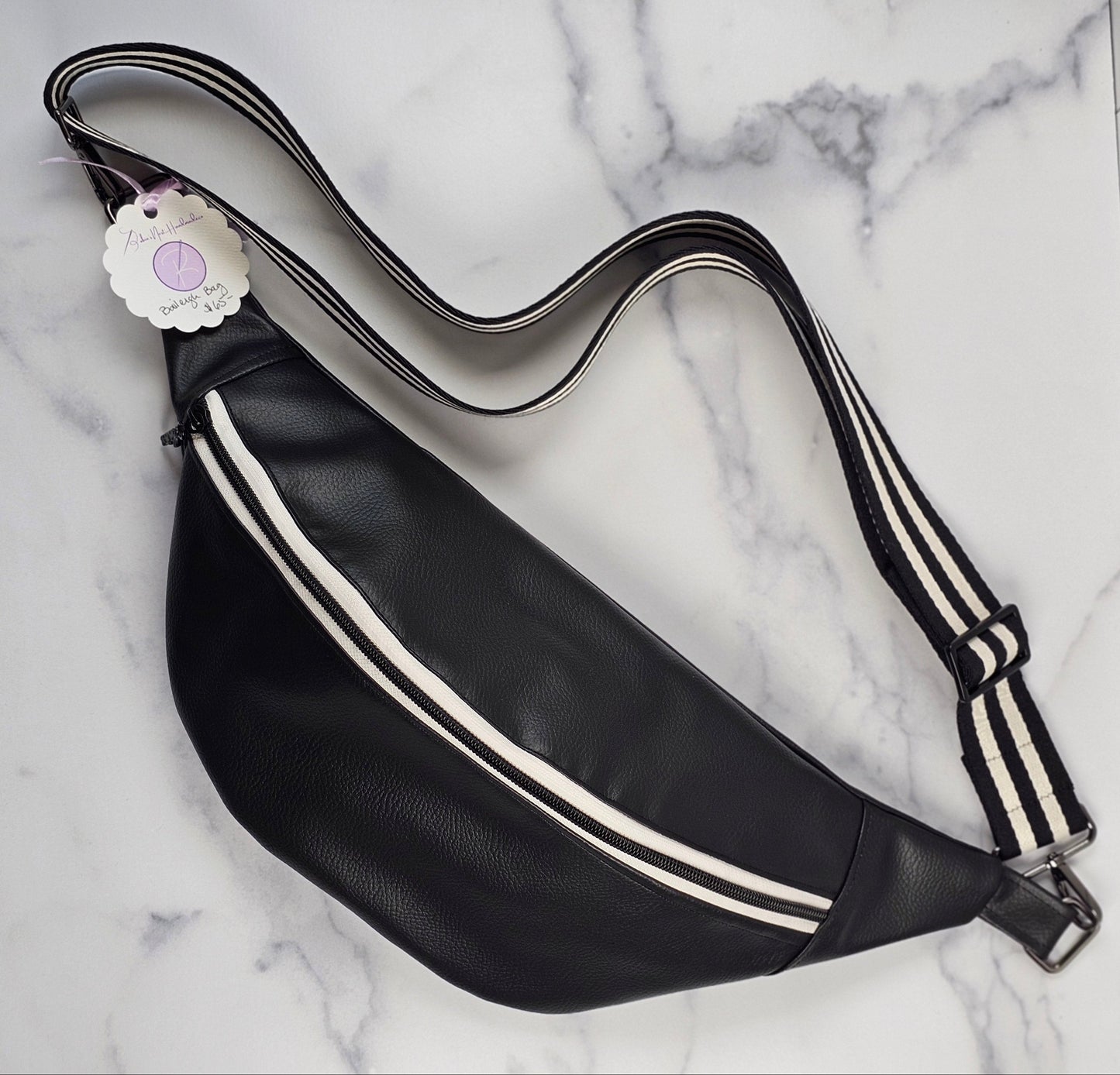 Black pebble faux leather sling, fanny pack with floral interior with one zipper pocket and one slip pocket. White zipper with "kindness" zipper pull and black and white striped strap.